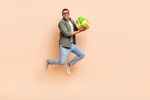 全长照片的酷开朗的家伙穿着卡其布衬衫跳高拿着礼物盒孤立的米色背景图片下载