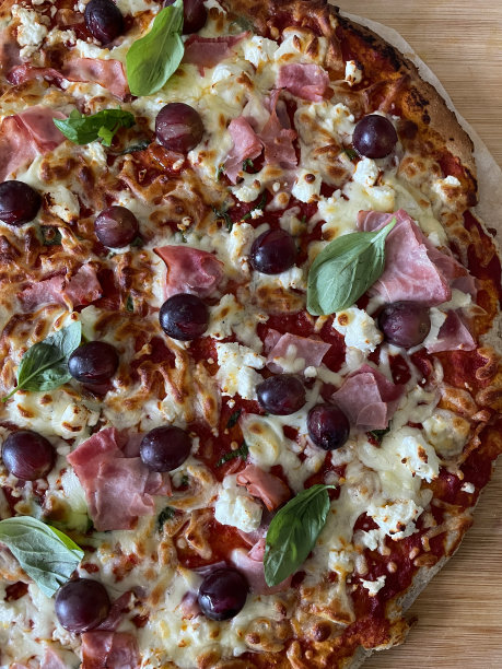 在木制砧板上新鲜出炉的自制披萨的特写图片，火腿、菲达奶酪、红葡萄、马苏里拉奶酪、罗勒叶浇头、丰富的番茄酱，高架视图图片下载