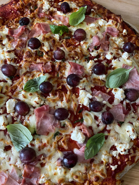 在木制砧板上新鲜出炉的自制披萨的特写图片，火腿、菲达奶酪、红葡萄、马苏里拉奶酪、罗勒叶浇头、丰富的番茄酱，高架视图图片下载