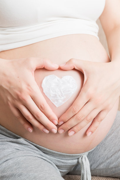 一个怀孕的女孩坐在家里的床上，在肚子上涂了一种抗妊娠纹霜。怀孕，做母亲，准备和期望的概念图片下载