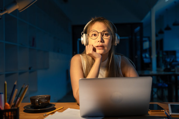 年轻的亚洲女子戴着白色耳机听音乐，晚上在笔记本电脑上做她的项目，试图为她的生意想出一个主意图片下载