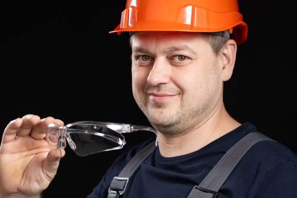 在黑色背景上，一个英俊的工人建筑工人穿着工作服，戴着头盔和眼镜。图片下载