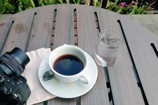 一个黑色的咖啡杯放在木桌上，相机和一杯水，背景是花园。图片下载
