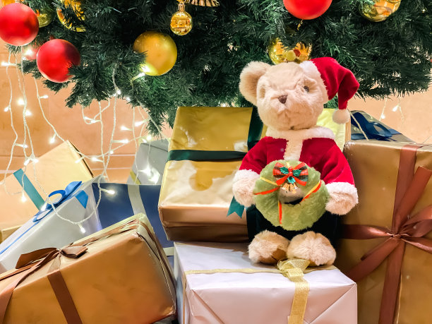 圣诞泰迪熊穿着节日服装，手拿圣诞花环，坐在圣诞树下包裹好的礼物上图片下载