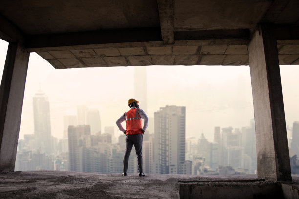 印度男工程师戴着头盔，穿着背心，站在建筑工地，眺望建筑物和城市景观。长镜头，广角镜头。副本的空间。房地产。图片下载