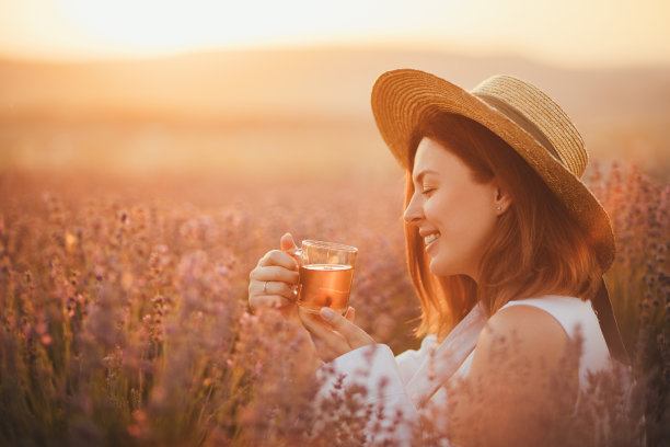 年轻快乐的女人喝着花草茶，坐在美丽的薰衣草地里看日落。图片下载