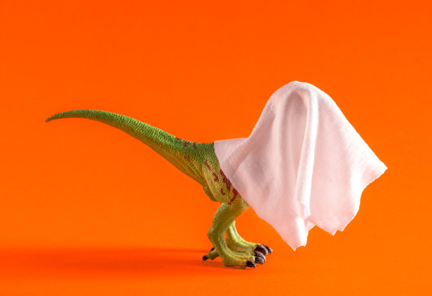 有趣的恐龙穿着白色床单作为幽灵服装在橙色背景。极简艺术海报。图片下载