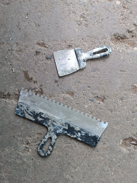 两个抹刀，用于在混凝土板上铺设瓷砖。建筑工具躺在地板上。脏的工作工具。宽抹刀和小抹刀，在旧的坑洼混凝土上涂上胶水。地板的概念。图片下载