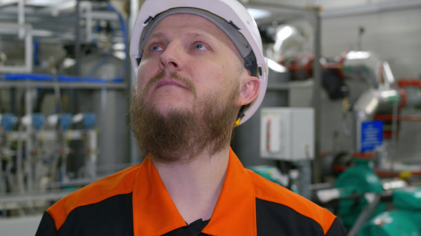 在一幢抽取石油和天然气凝析液的工业生产车间里，一名戴着白色头盔、留着胡子的工程师的脸部特写。石化行业图片下载