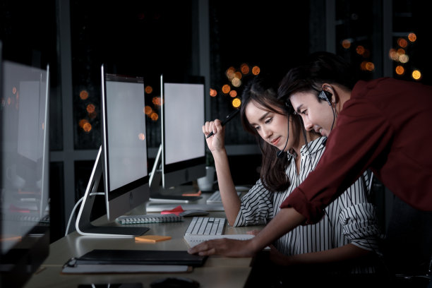 快乐的亚洲男女在呼叫中心咨询台与队友讨论，客服主管团队使用麦克风耳机在深夜办公室支持客户图片下载
