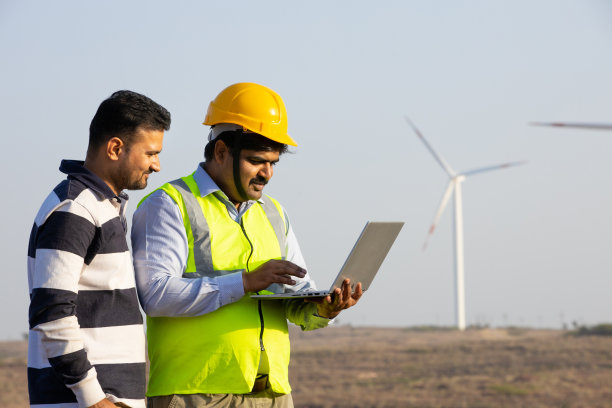 印度工程师使用笔记本电脑，展示控制风车农场发电的操作，亚洲人戴着黄色头盔和背心，在风力涡轮机农场工作，清洁和绿色能源。图片下载