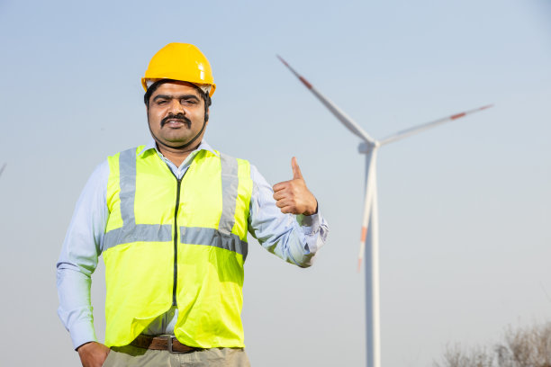 自信的印度工程师戴着黄色头盔，穿着背心，站在风车农场或风力涡轮机农场，清洁和绿色能源。运行发电;图片下载