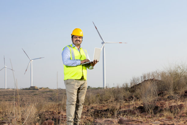 印度工程师用笔记本电脑控制和维护风车农场的运作来发电，亚洲人戴着黄色头盔和背心在风力涡轮机农场工作，清洁和绿色能源。图片下载