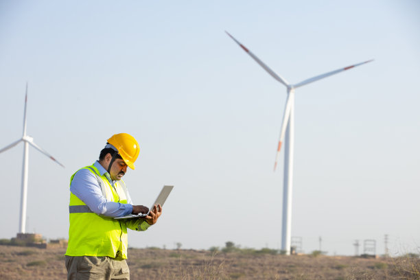 印度工程师用笔记本电脑控制和维护风车农场的运作来发电，亚洲人戴着黄色头盔和背心在风力涡轮机农场工作，清洁和绿色能源。图片下载