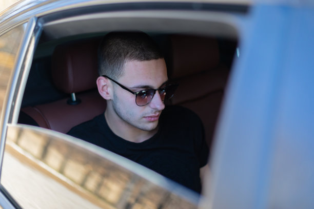 高清照片，一个年轻人坐在汽车的后座上用自己的手机在路上，制定计划，搜索，上网和媒体，聊天，微笑，享受公路旅行在一个美丽的阳光明媚的日子。图片下载