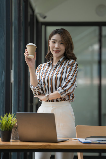 年轻自信的亚洲女商人端着咖啡杯站在办公室里。图片下载