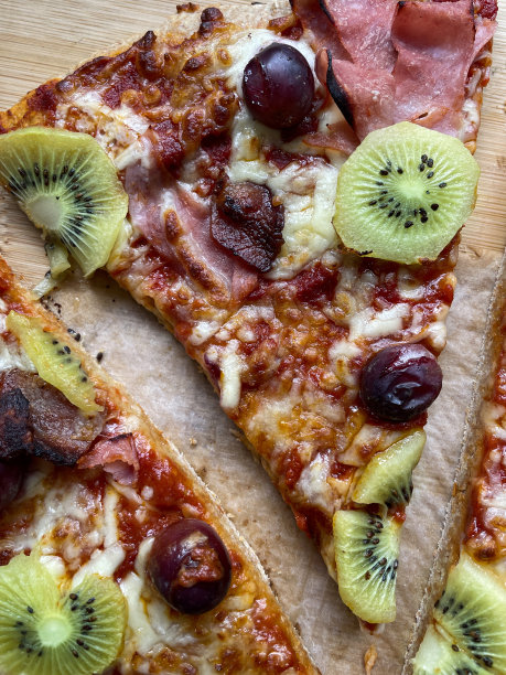 新鲜出炉的、三角形切片的、自制披萨、红葡萄、火腿、猕猴桃片、磨碎的马苏里拉奶酪浇头、丰富的番茄酱、防油羊皮纸背景、高架视图的特写图像图片下载