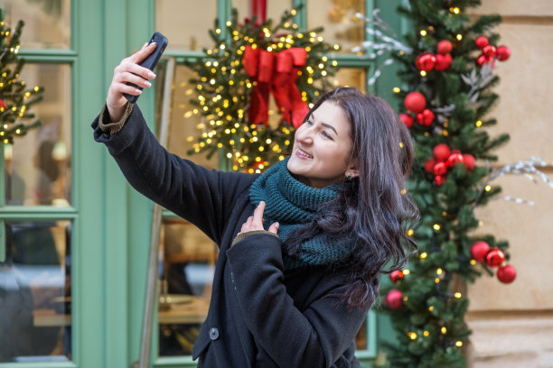 快乐的女人在城市里使用智能手机，背景是节日的圣诞装饰。图片下载