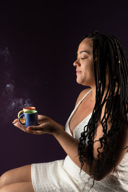 一个漂亮女人的画像，头发上扎着辫子，坐着，穿着白色的衣服，两杯冒着热气的咖啡。工作室拍摄深紫丁香背景。积极的人。图片下载