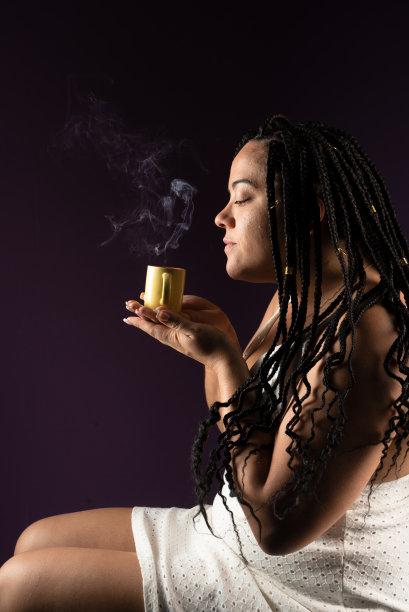 一个美丽的女人的画像，头发上扎着辫子，坐着，穿着白色的衣服，端着一杯热气腾腾的咖啡。工作室拍摄深紫丁香背景。积极的人。图片下载