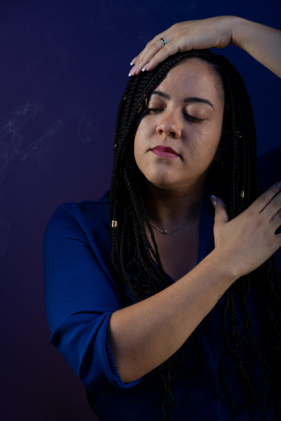 一个美丽的女人，扎着辫子，穿着蓝色的衣服，抚摸着她的头和胸部。微笑和快乐。孤立的深蓝色背景。图片下载