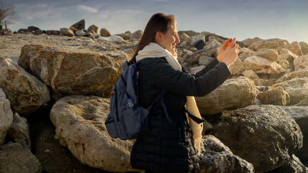 一名年轻女子站在岩石上用智能手机拍照，捕捉到了冬天大海的美丽。适合度假和旅行。图片下载
