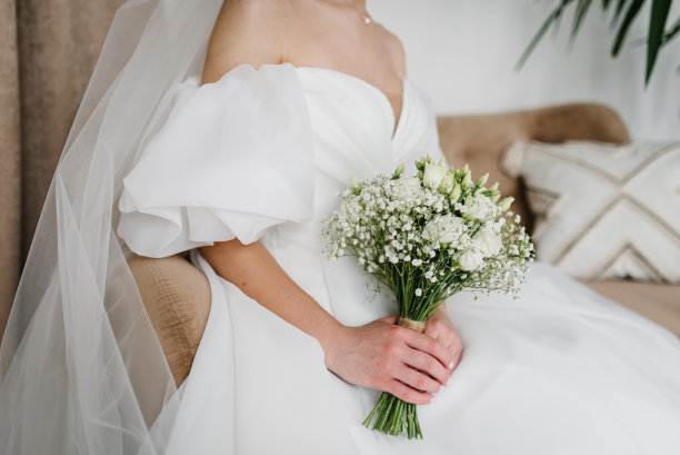 一位迷人的新娘穿着时髦的礼服，在家里拿着一束美丽的白玫瑰和吉卜赛花。新娘手中的婚礼花束。早上准备。准备结婚的女人。特写镜头图片下载