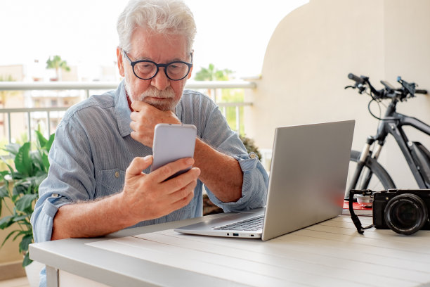 严肃的白发老人戴着眼镜使用手机，老年男性坐在户外的家庭露台上使用现代技术的笔记本电脑图片下载
