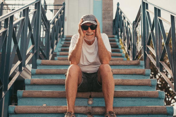 一位戴着帽子和太阳镜的高级英俊男子的肖像，看着相机微笑，坐在户外楼梯上放松。享受假期或退休生活的无忧无虑的老人图片下载