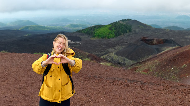 徒步旅行在欧洲大陆最高的火山-埃特纳。年轻的女人微笑着，穿着黄色的雨衣，在一个有风的日子里，在埃特纳火山全景的山顶用手做心形。图片下载