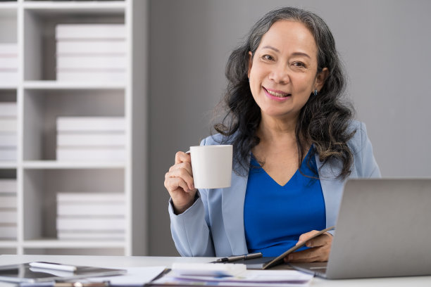 年长成熟的女人快乐年轻地坐在舒适的办公椅上，享受她的早晨咖啡。图片下载