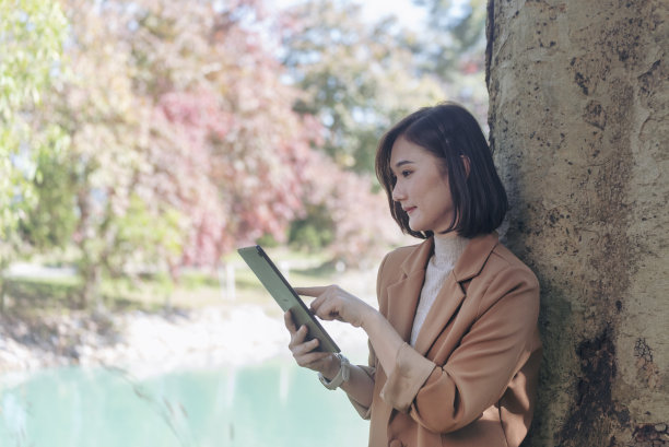 在秋天的绿色公园户外，一名亚洲妇女手持数码平板电脑在网上购物。女性用手触摸指向智能平板电脑，使用移动应用程序工作旅行。漂亮的亚洲女人，智能科技图片下载