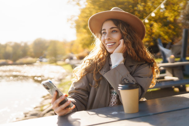 戴着帽子的年轻女游客在湖边的长椅上喝着咖啡，享受着阳光明媚的天气。图片下载