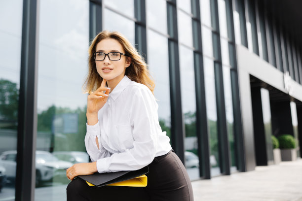 优雅自信的年轻女子，穿着正式，站在户外，以商务中心为背景，拿着一个装有文件的文件夹，看着相机图片下载