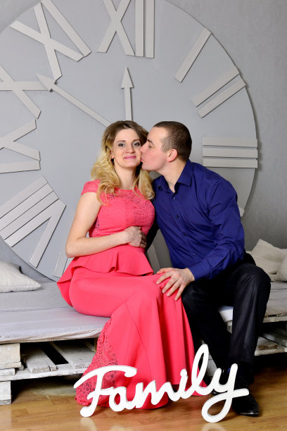 一对迷人的年轻夫妇正在家里庆祝节日。站在美丽的圣诞树背景上，丈夫拥抱着怀孕的妻子。一个男人和他怀孕的妻子站在时钟旁。墙上的新年钟。高质量照片图片下载