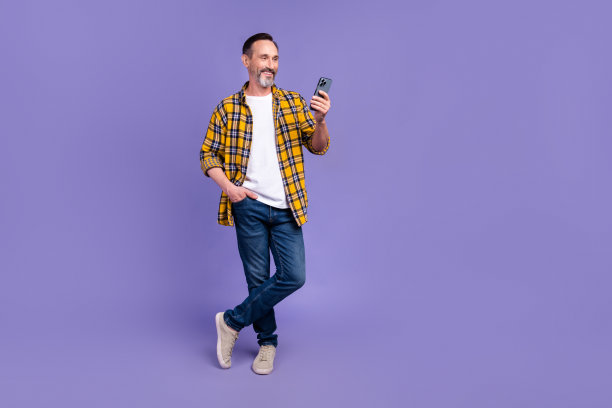 酷男人的全尺寸照片穿黄色格子衬衫牛仔裤裤子看着智能手机手臂在口袋里孤立的紫色背景图片下载