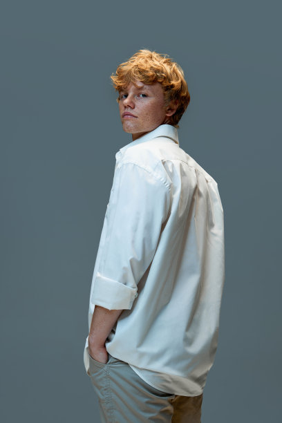 严肃的年轻红卷发男子的肖像，穿着白衬衫，站在灰色的背景下看着相机图片下载