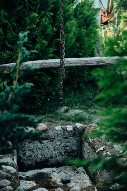一个生锈的旧钩子挂在一个石头井上，周围是彩色玻璃窗和绿色植物图片下载