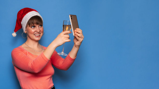 一位戴着圣诞老人帽、面带微笑的迷人女子拿着一杯香槟和一部手机，背景是明亮的蓝色。圣诞节的气氛在大流行。放文字的地方。图片下载
