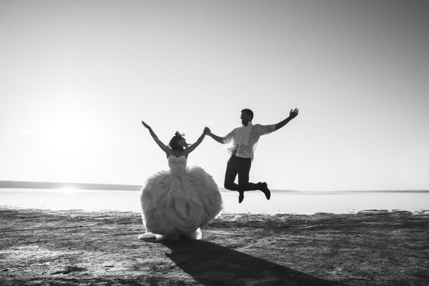 新婚夫妇正在海滩上蹦蹦跳跳。阳光明媚的夏日照片。新娘披散着头发，穿着露肩裙。海洋仪式。海边的爱情故事。沙子，水和地平线。黑色和白色。图片下载