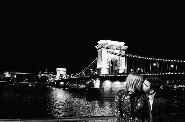 一对热恋情侣在布达佩斯吊桥上亲吻拥抱。欧洲城市夜晚的城市灯光。男人和女人的剪影附近的河全景。时尚的黑白时尚照片图片下载