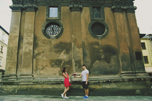 一对相爱的情侣走在古老的古镇里。在中世纪的石头教堂附近，穿短裤的男子正在给他穿粉色连衣裙的女孩送花。浪漫的夏天时髦的照片。害羞的情绪。图片下载