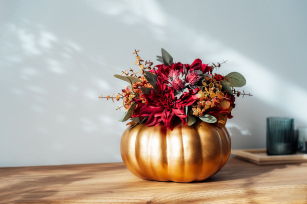 秋天的花束明亮的人造花在一个金色的南瓜花瓶，蜡烛在木制控制台与白色的墙壁与阴影的背景。Hygge家居秋季装饰，营造舒适的家居氛围。图片下载
