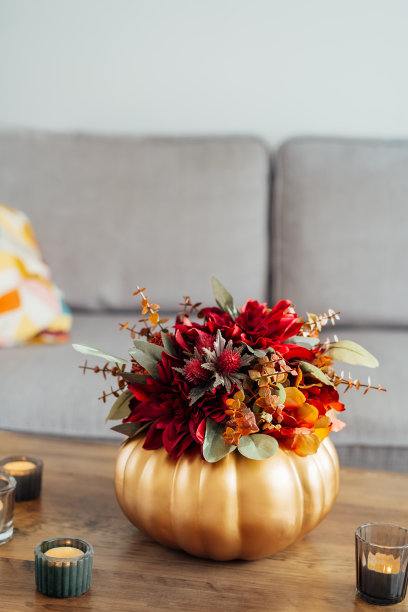 秋天的花束明亮的人造花在一个金色的南瓜花瓶里，燃烧的蜡烛在客厅室内的木制咖啡桌上。Hygge家居秋季装饰，营造舒适的家居氛围。垂直图片下载