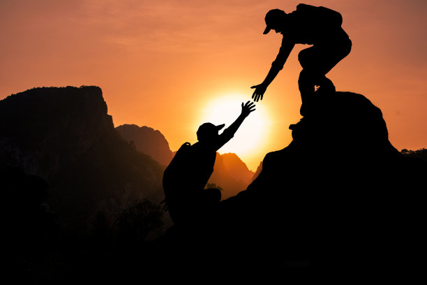 在阳光的映照下，登山者伸出援助之手把他的朋友拉上山顶。图片下载