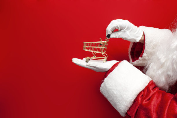 圣诞老人穿着西装，戴着手套，捧着红色背景上的空购物车，手里站着空的小推车图片下载