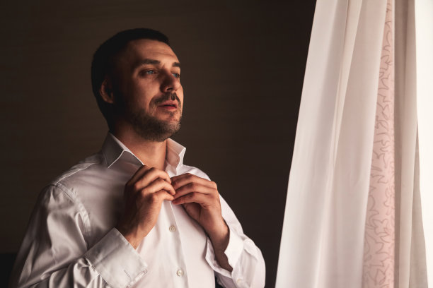 自信的新郎在酒店房间扣上白衬衫，严肃地看着窗户。年轻人正在梳妆准备迎接新娘。年轻家庭观念，美满婚姻。复制广告文本空间图片下载