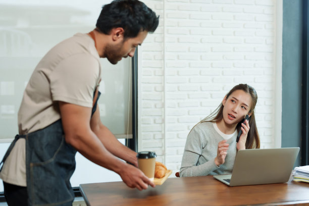 漂亮的亚洲女人坐在桌边在咖啡馆工作。印度男服务员端来咖啡。牛角面包放在木托盘上，端上餐桌。小生意咖啡店的服务员图片下载