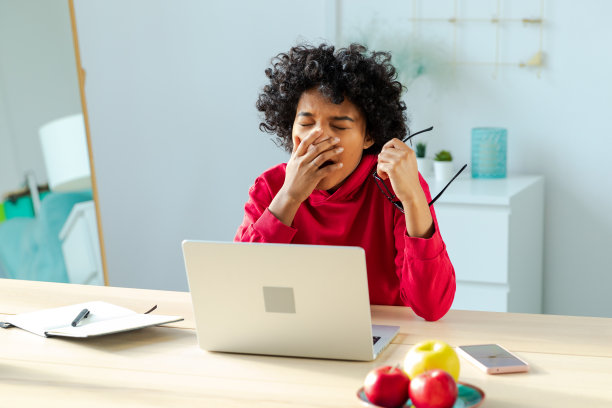 昏昏欲睡的非裔美国女人坐在桌子旁，拿着笔记本电脑，打着哈欠。劳累过度的女孩懒于工作。学生女商人下班后在家里办公室用笔记本电脑放松。图片下载