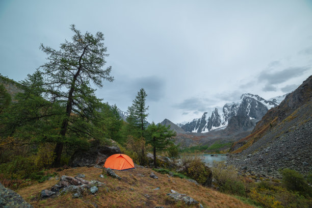 戏剧性的景观，在岩石和秋天的植物群中，在森林山上有一个橙色的帐篷，在多云的天空下，可以看到大的雪山。高高的山上，孤独的帐篷和褪色的秋色。图片下载